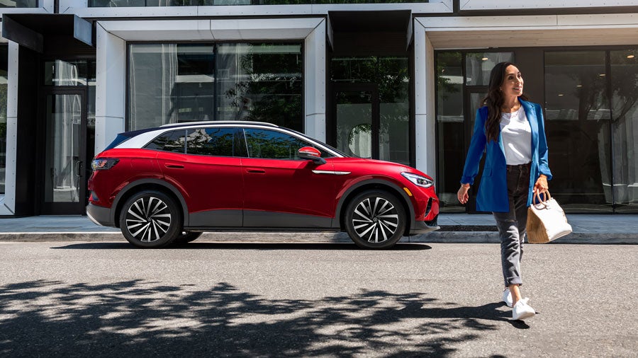 A woman walks away from an Aurora Red Metallic ID.4 SUV that’s parked on an urban street in front of a modern office building.